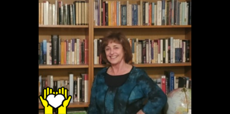 Lynn Kennedy poses for a photo in front of a book shelf. A yellow graphic of hands holding a white heart outlined in black in the bottom left corner