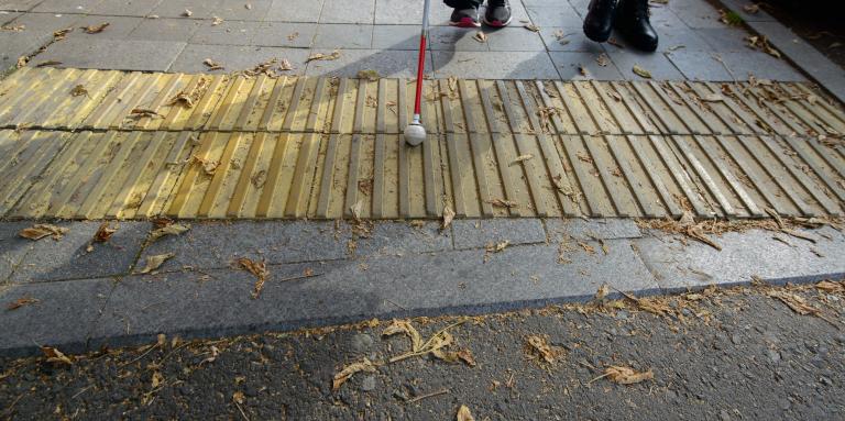 A white cane with a rolling ball tip scans tactile paving on a sidewalk.
