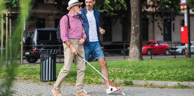 Two men take a walk along a paved path in a grassy area. One man navigates with a white cane.