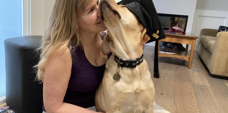 Laurie sits on her living room floor with her guide dog, Bridget, a yellow Labrador retriever-golden retriever cross. Bridget wears a graduation cap and sits in Laurie’s lap. Laurie affectionately presses her face against the Bridget’s fluffly face. 
