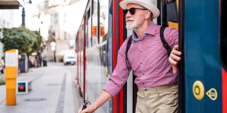 An image of an older man getting out of a streetcar and exiting onto a busy street. He uses a white cane, and is wearing a red and blue collared shirt, khakis and a fedora. He is smiling.