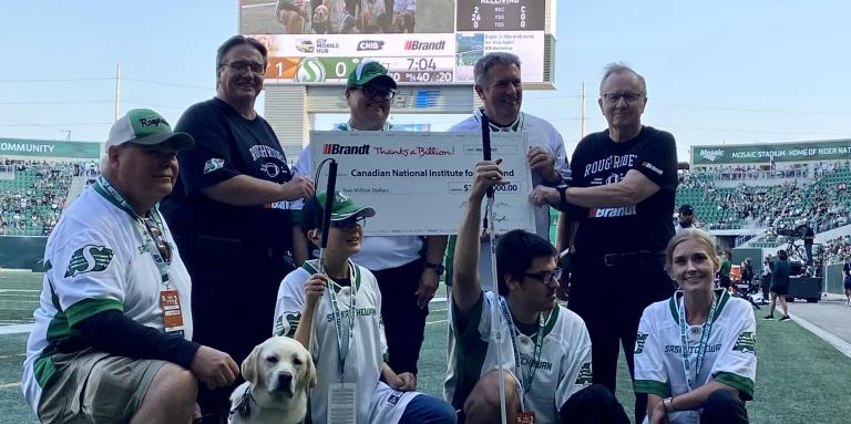 Representatives from CNIB and the Brandt Group of Companies pose together for a photo while holding the $2M cheque at the Saskatchewan Roughriders Game. There are two CNIB Guide Dogs wearing green jerseys. 