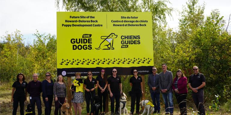 A group of 13 people, including CNIB Guide Dogs staff and municipal officials from the Town of Georgina, gather outdoors in rural Georgina to unveil signage for the future home of the Howard & Delores Beck Puppy Development Centre.