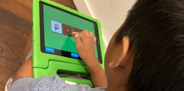 A young boy plays on a tablet. He is sitting on the ground and the tablet is in his lap is in a protective green rubber case. The picture is an overhead view of the child tapping the tablet screen and interacting with the educational app. 