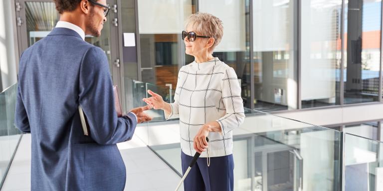 Two employees engaged in discussion in a brightly lit office space. One of the employees, a middle-aged woman with short, grey hair, uses a white cane