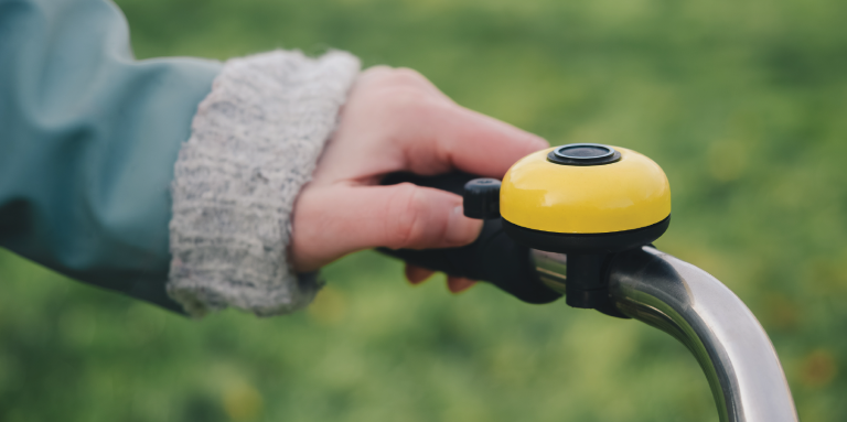 A person’s hand rings a yellow bicycle bell. The bell is mounted on the bicycle handlebar. 