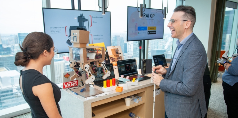 A man in a suit holding a white can speaking to a woman in front of a table full of robotic tech