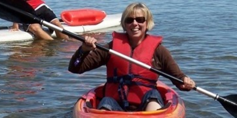 Joanne smiles and poses in an orange kayak for a photo on the lake. She’s wearing sunglasses, a red life jacket, and holding her paddle. 