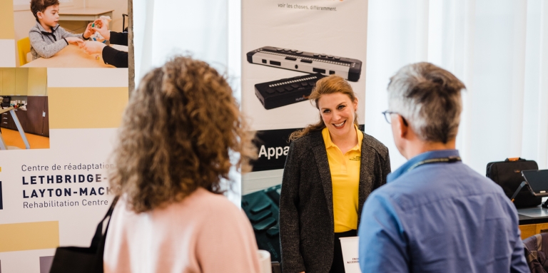 A woman behind an exhibit table speaking to a man and woman.