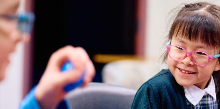 Two schoolchildren in a classroom laughing. One is holding a small blue ball. 