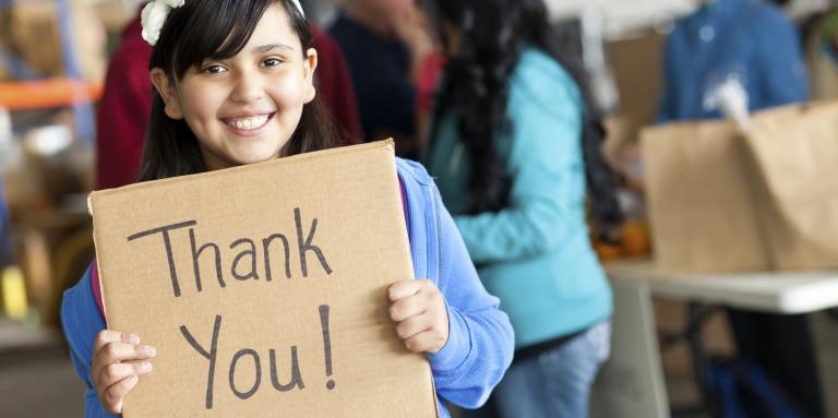 A young girl smiles and holds a cardboard sign that reads 'thank you!' 