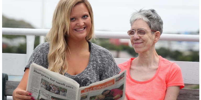 A young woman reads a newspaper to an older woman.