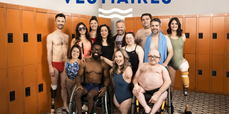 A group of people, some of whom have visible disabilities, wear swimwear and pose together in a locker room