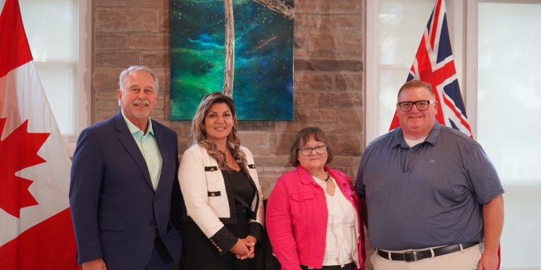 The Honourable Neil Lumsden Ontario’s Minister of Sport, Angela Bonfanti, President & CEO for CNIB, Sharon Cadeau, Ontario Vice Chair of Métis Nation of Ontario, and the Honourable Graydon Smith, MPP for Parry Sound–Muskoka stand in front of the fireplace at CNIB Lake Joe. The Canada flag and Ontario flags are beside us. 