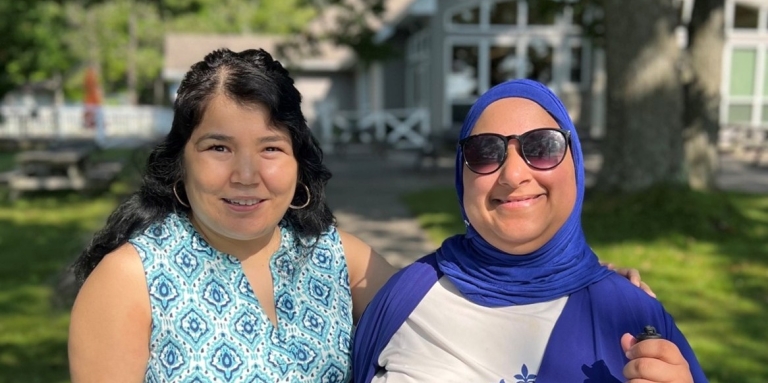 Najla [left] and Salma [right] smile outdoors in front of a leafy tree and a light grey lodge-style building at CNIB Lake Joe.