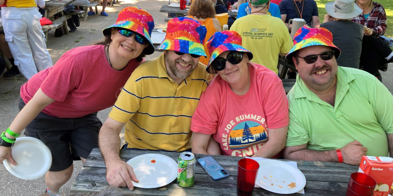 Four people eat lunch outdoors wearing bright t-shirts and bucket hats 