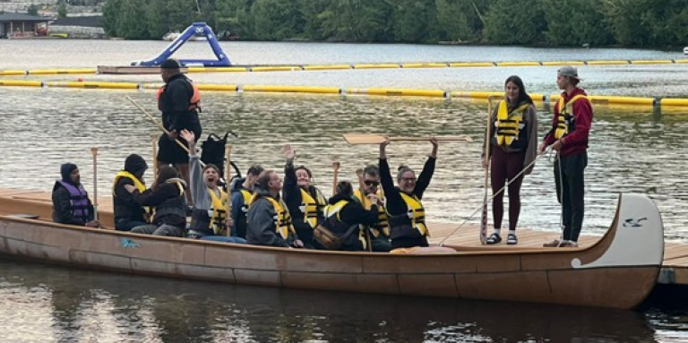 A group of people in a big Voyageur canoe at the dock. They are wearing yellow lifejackets and have their hands raised in the air.