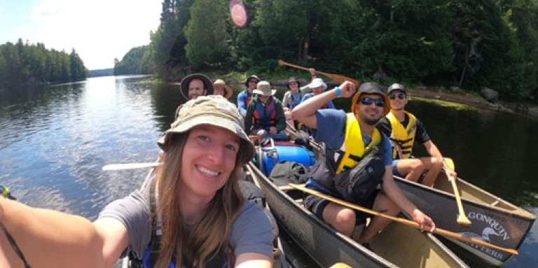 A cheerful group of CNIB Lake Joe participants paddle through a calm lake surrounded by lush green forest in Algonquin Park. 