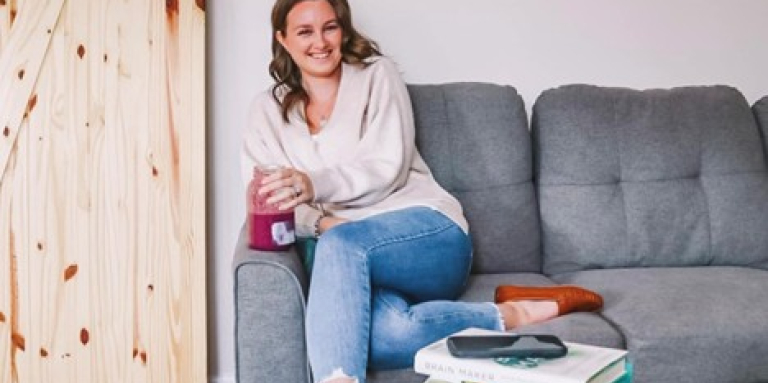 A young woman sitting on a gray couch wearing jeans and a cream coloured sweater. There are books about health stacked on a coffee table in front of her. She is smiling, holding a glass jar filled with fruit juice.