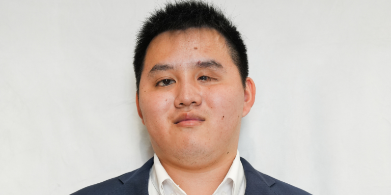 Zhicheng, a young Asian man wearing a navy suit with a white dress shirt, stands in-front of a white background. Zhicheng is smiling and the photograph resembles a professional headshot. 