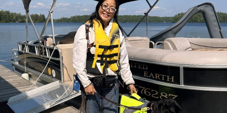 Kathryn is standing on the dock with a pontoon boat behind her. She is wearing a lifejacket and holding her guide dog on harness. It's a bright sunny day and she is smiling at the camera.