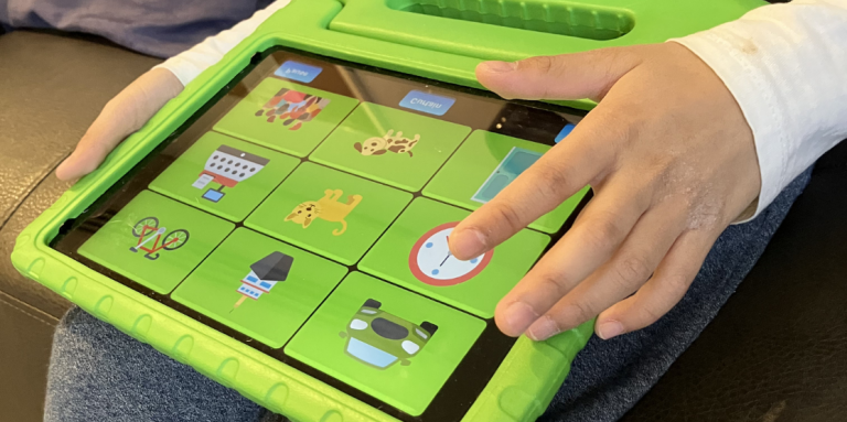 A child’s hands hold an iPad in a bright green protective case, tapping a grid of colourful icons including animals, objects, and a clock on the screen.