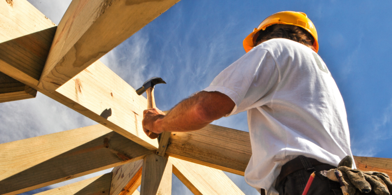Construction worker wearing a hard hat uses a hammer to secure wooden roof beams, viewed from below against a bright blue sky.