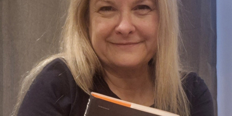 Mary Ellen is seated while hugging a hardcover book. She is smiling at the camera.