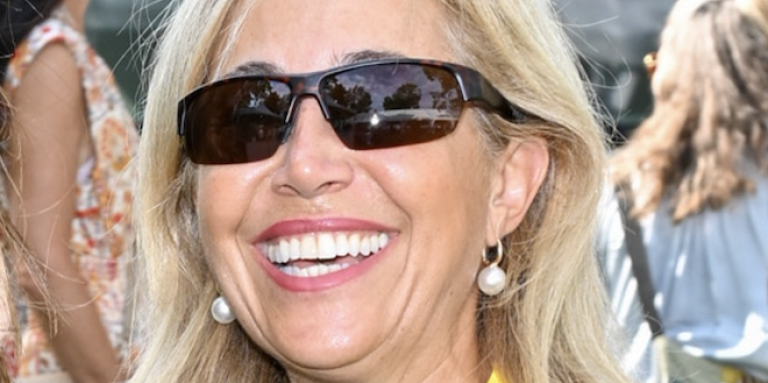 Frances is outdoors with a group of people milling about behind her. She is wearing dark glasses and smiling at the camera.