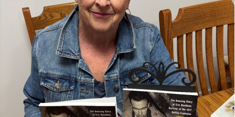 Author Marilyn is sitting at a dining room table holding up copies of her book, “The Blind Mechanic.”