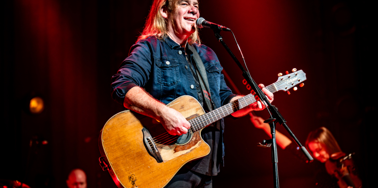 Musician Alan Doyle stands on stage in front of a microphone holding a guitar 