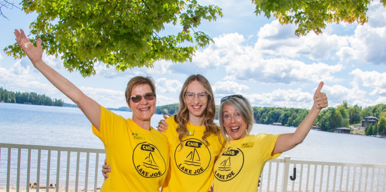 Monique Pilkington, Emma Van Dyk, and Sherri Helsdingen stand together by the Lake Joe waterfront, wearing yellow Lake Joe T-shirts with their arms around one another. They smile at the camera and give thumbs-up gestures.