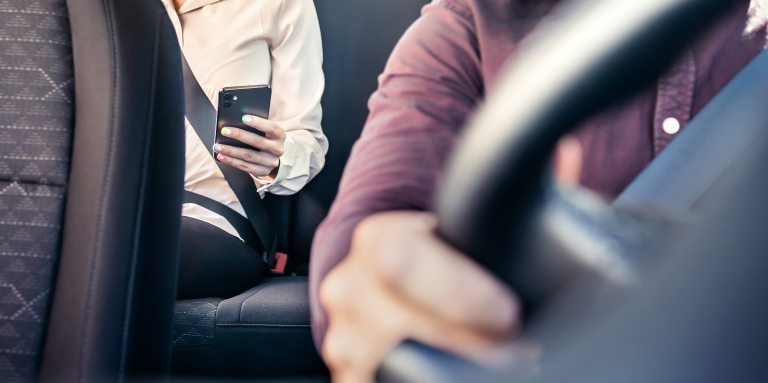 A passenger wearing a seatbelt sits in the back seat of a rideshare car, holding a smartphone while the driver steers from the front seat.
