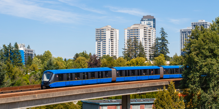 A Vancouver SkyTrain car crosses an elevated track above a tree-filled neighbourhood with residential high-rise buildings under a clear blue sky.