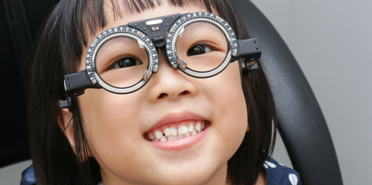 A smiling young girl wears a trial frame with adjustable lenses during an eye exam.