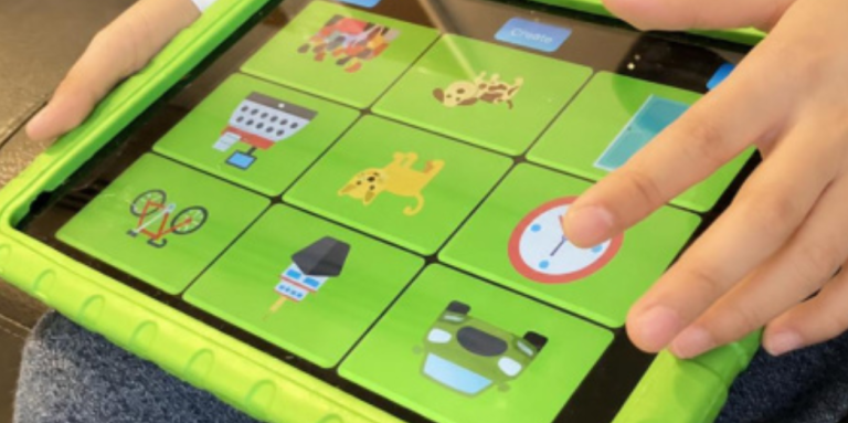 A child’s hands hold an iPad in a bright green protective case, tapping a grid of colourful icons including animals, objects, and a clock on the screen.