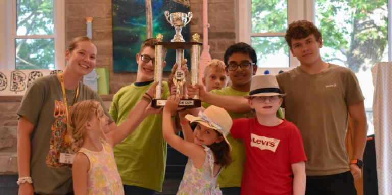 Seven children and youth gather around a large trophy in lounge at CNIB Lake Joe, smiling as they hold it up together. The trophy features a silver cup above three tall gold columns.