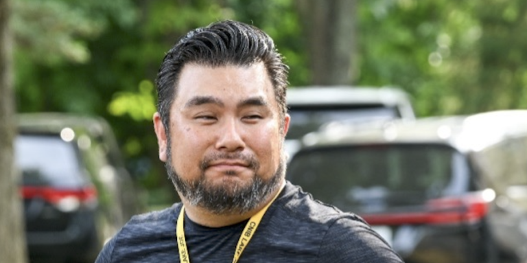 Eugene Chong stands outdoors with his hands on his hips and a slight smile. He has short, swept-back dark hair and a neatly trimmed salt-and-pepper beard. He wears a dark athletic T-shirt, a yellow CNIB Lake Joe lanyard with name badges, and a black wristwatch. Sunlit green trees and parked vehicles are softly blurred behind him.