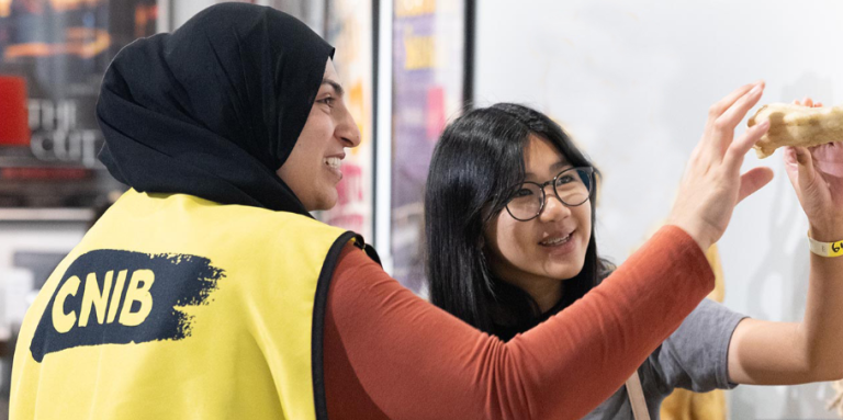 Two women, one wearing a CNIB vest, smile while holding and examining an object together indoors.