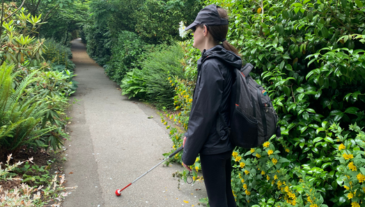 Abby walks along a lush green nature pathway and navigates with their white cane. Abby is turned away from camera.