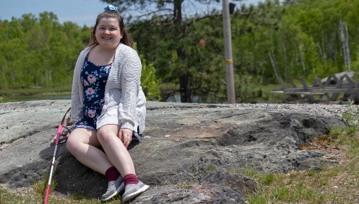Alicia smiles and sits outdoors on a large rock. Behind her are trees. She is wearing a summer floral dress. Her white cane rests on the rock to the right of her.