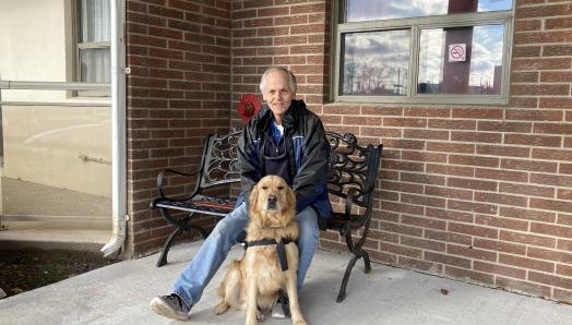 Photo of Terry sitting outside on a metal bench, smiling for the camera with Bert sitting front of him. 