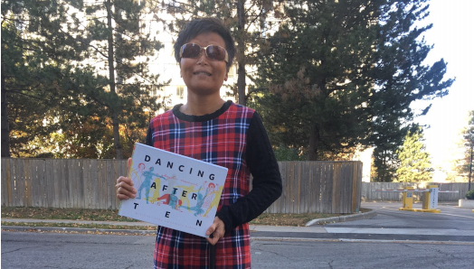 Vivian Chong stands outdoors and holds her book, Dancing after TEN. Her black guide dog, Catcher, stands at her feet. Vivian is wearing a plaid dress, sunglasses, and a big smile. 