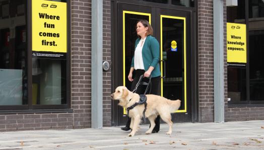 A woman smiling while walking with her golden retriever guide dog along a sidewalk in front of a CNIB Community Hub.