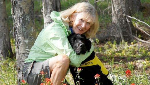 Cheryl kneeling next to Irwin, a black Labrador retriever wearing a yellow Future Guide Dog vest, in a forest in Canmore Alberta surrounded by trees and greenery.