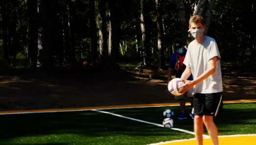 A teenaged camper wearing face mask preparing to kick a soccer ball on the new soccer pitch at CNIB Lake Joe 