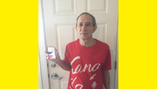 Bill Chadd pose pour une photo avec son téléphone intelligent. Il porte un tee-shirt rouge du Canada et se tient devant une porte en souriant et en tenant un iPhone dans la main droite.