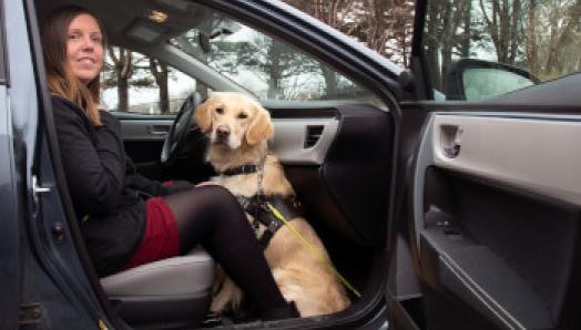 A woman is sitting in the front passenger seat of a car with the door open; her guide dog, a golden retriever, is sitting at her feet in the passenger footwell.