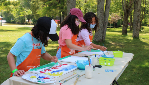 Three girls painting at a crafts table set up outside on the lawn.