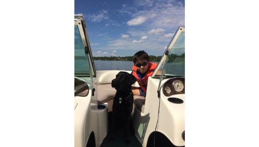 Keaton est assis sur un bateau dans l'eau avec son bras autour d'Indy, un labrador golden-retriever noir, qui est assis sur le bateau à côté de lui.